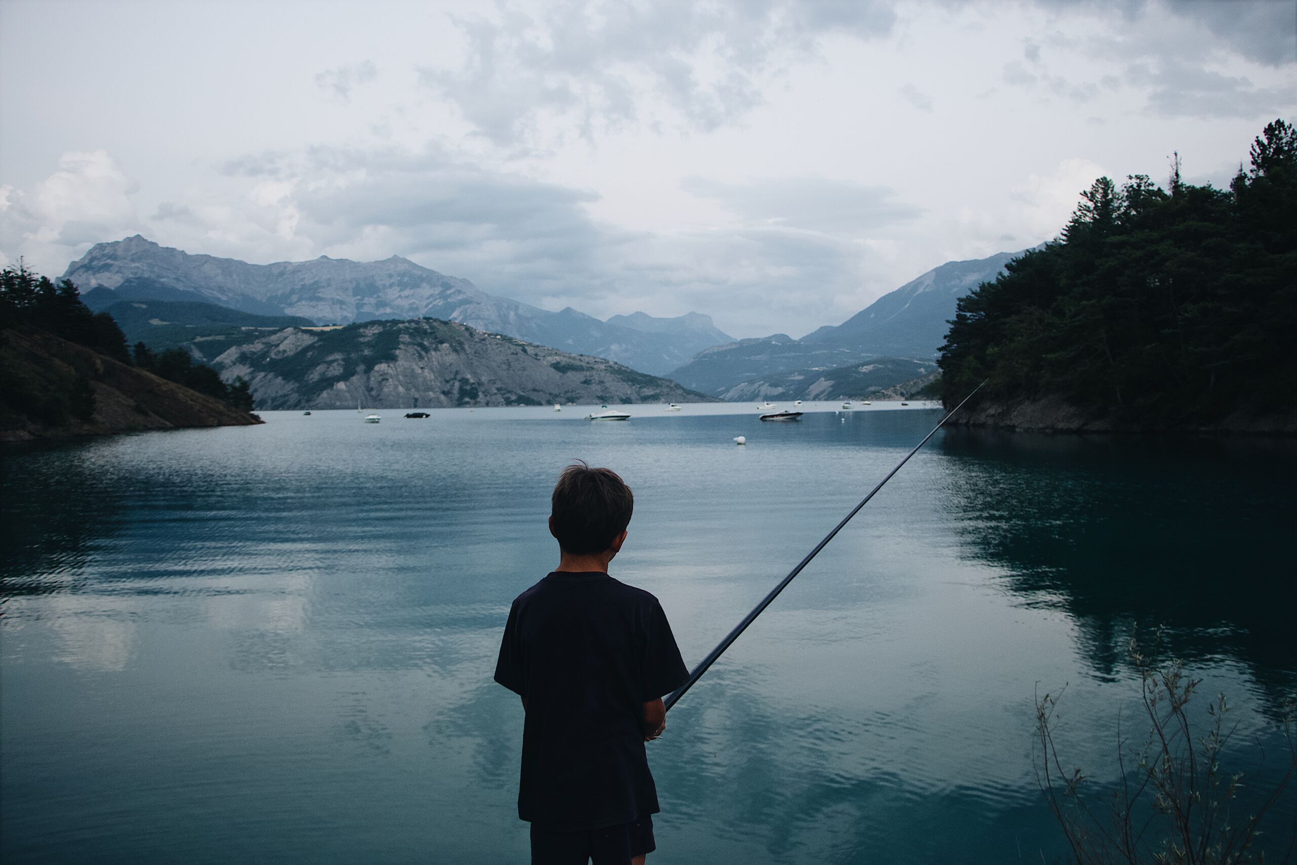 A boy fishing in the water with mountains behind him.